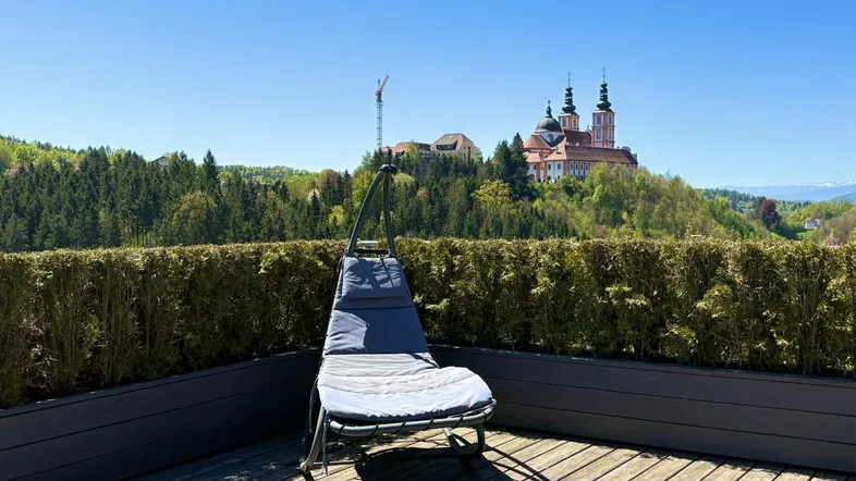 Sonniger Terrassenbereich mit Holzbelag, Liegestuhl und weitem Blick auf eine historische Kirche und grüne Hügel.