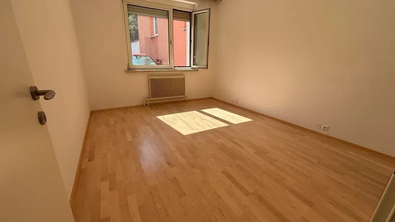 Helles, leeres Zimmer mit Holzboden und Fenster, das viel Tageslicht hereinlässt.