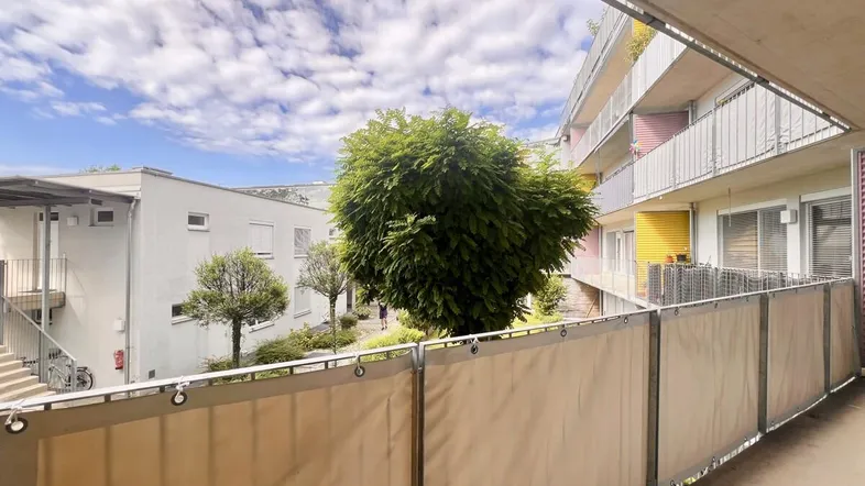 Blick vom Balkon auf den begrünten Innenhof mit weiteren Wohngebäuden und blauem Himmel.