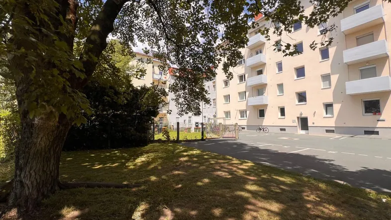 Mehrfamilienhaus mit heller Fassade, umgeben von Grünflächen und Bäumen, mit Parkmöglichkeiten.