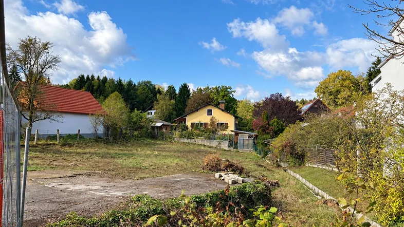 Leeres Grundstück mit umliegenden Häusern und Bäumen unter blauem Himmel.