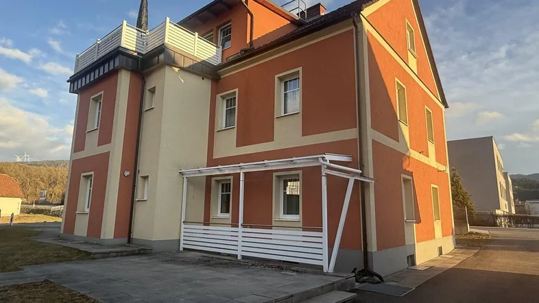 Mehrstöckiges Gebäude mit orangefarbener Fassade, einer Terrasse im Erdgeschoss und einer Dachterrasse.