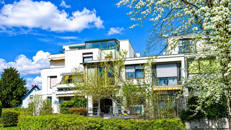 Modernes Wohngebäude mit viel Grün und blühenden Bäumen unter strahlend blauem Himmel.
