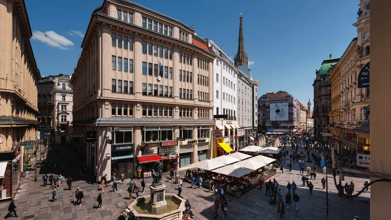 Eine belebte Einkaufsstraße in einer historischen Stadt mit vielen Passanten und Geschäften unter blauem Himmel.