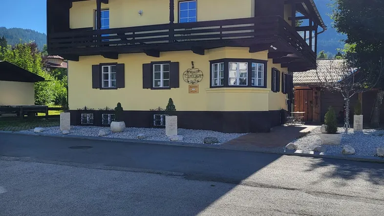 Sonnenbeschienenes gelbes Haus mit Holzbalkonen und grünem Dach in alpiner Umgebung.