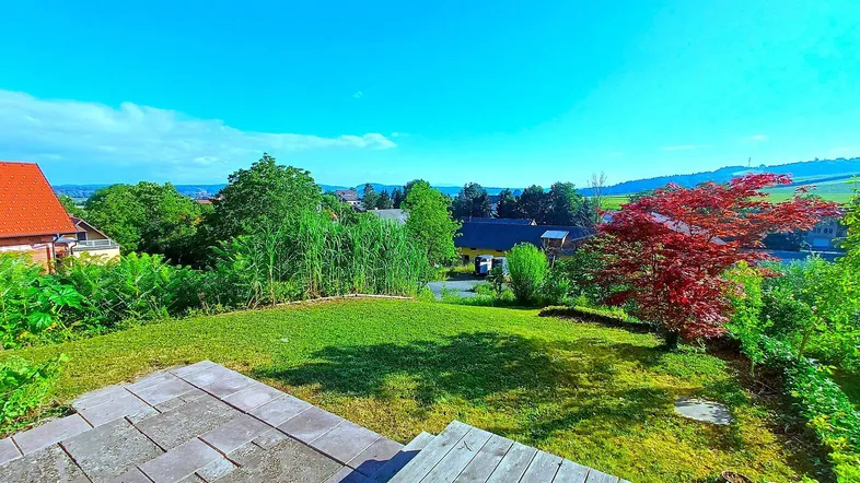Weitläufiger Gartenbereich mit Holzterrasse und Blick auf die grüne Landschaft unter blauem Himmel.
