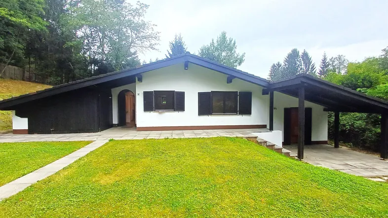 Einfamilienhaus mit weißer Fassade, großem Rasen und Carport, umgeben von Bäumen.