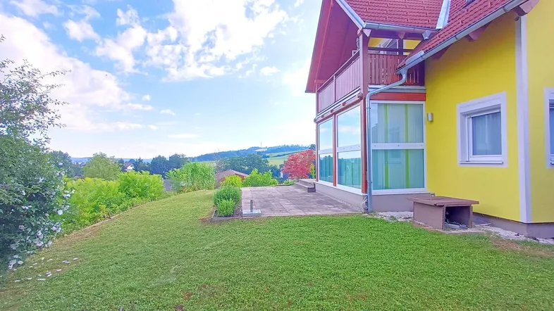 Gelbes Einfamilienhaus mit rotem Dach, Terrasse und Balkon, umgeben von grüner Natur.