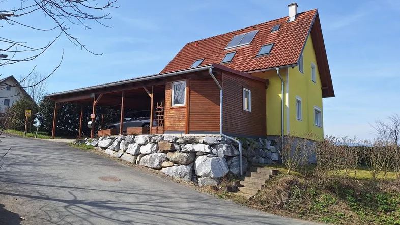 Gelbes Einfamilienhaus mit rotem Ziegeldach, Carport und Steinfundament an einem Hang.