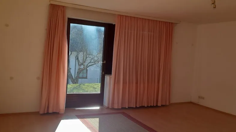 Heller Wohnraum mit großen Fenstertüren, rosa Vorhängen und Zugang zum Außenbereich.