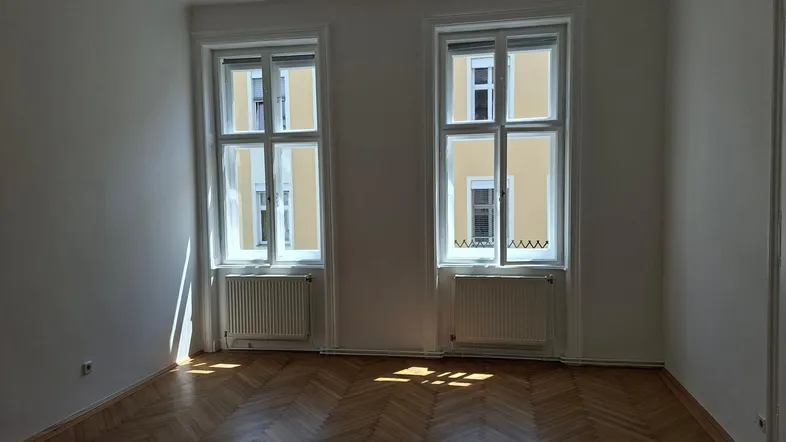 Helles Zimmer mit Fischgrätparkettboden und zwei großen Fenstern, die viel Tageslicht hereinlassen.
