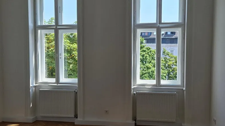 Helles Zimmer mit Fischgrätparkettboden, weißen Wänden und Blick ins Grüne durch große Fenster.
