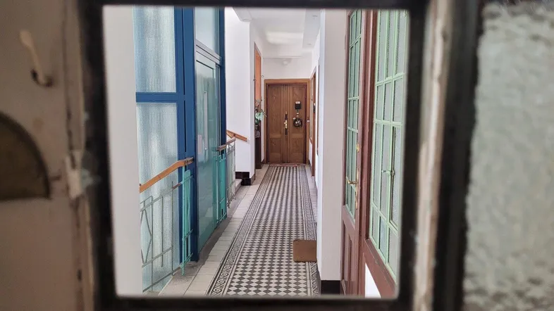 Blick durch eine historische Glastür in einen langen Flur mit gemusterten Fliesen und Holztüren.