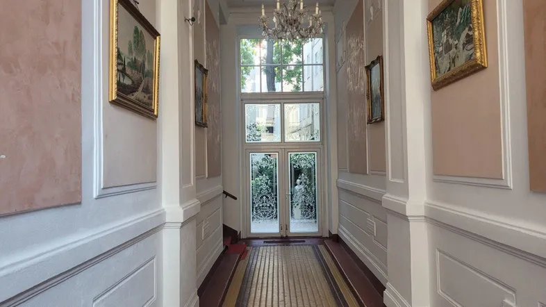 Eleganter Flur mit hohen Decken, Kronleuchter und historischen Gemälden, der zu einem großen Fenster führt.