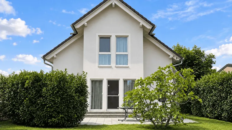 Weißes Einfamilienhaus mit gepflegtem Garten, kleiner Terrasse und grünen Hecken.