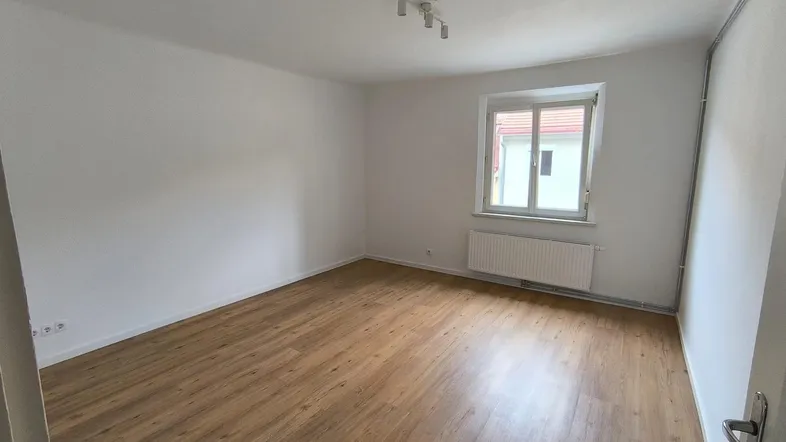 Helles, leeres Zimmer mit Laminatboden, weißen Wänden und einem Fenster mit Blick nach draußen.