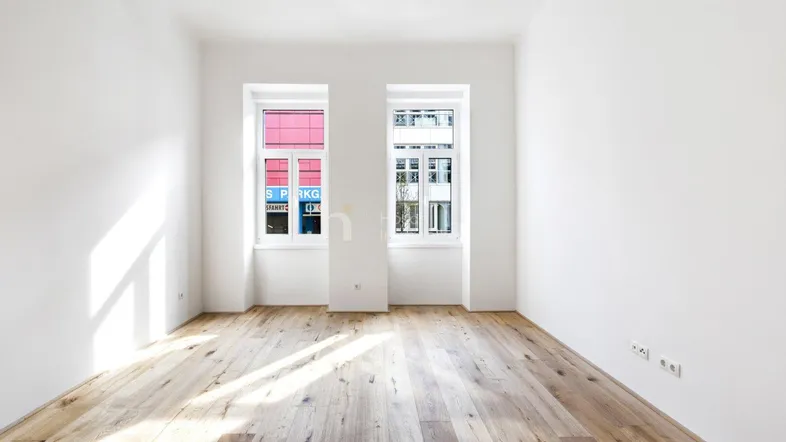 Helles, leeres Zimmer mit Holzboden und zwei Fenstern, das viel Tageslicht hereinlässt.