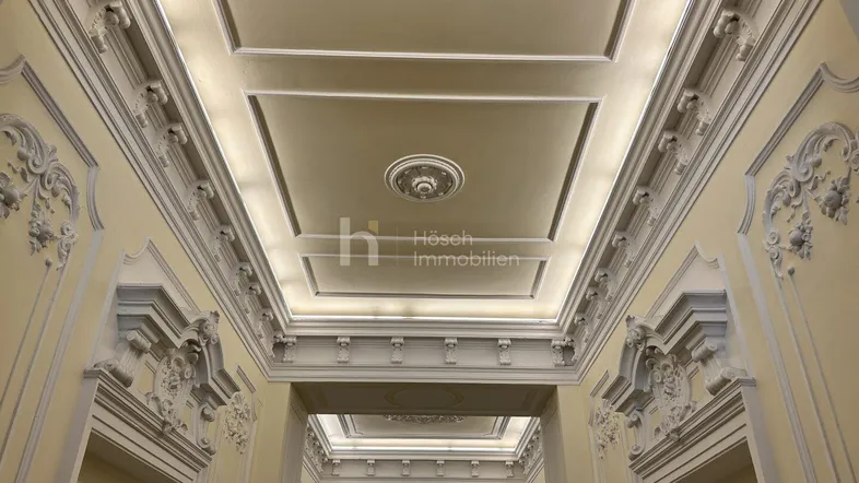 Eindrucksvoller Eingangsbereich mit aufwendigen Stuckverzierungen an Decke und Wänden, beleuchtet durch indirektes Licht.