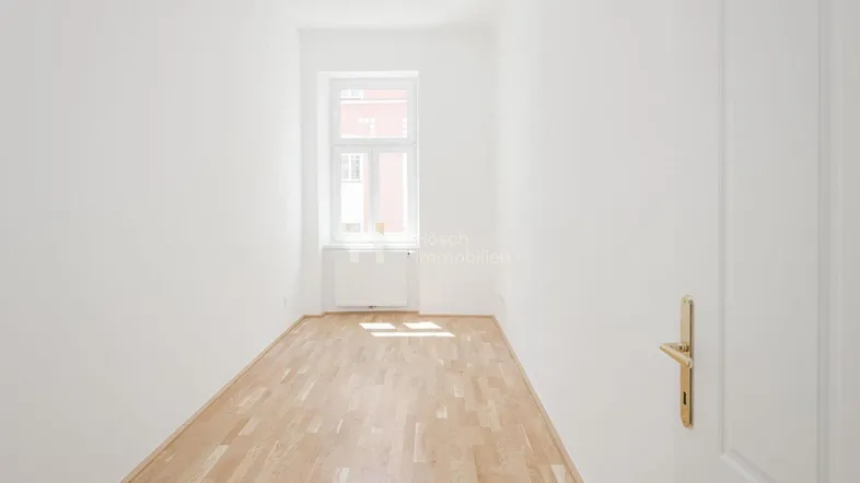 Helles, leeres Zimmer mit Parkettboden und einem Fenster, das viel Tageslicht hereinlässt.