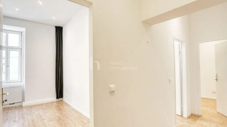 Heller Flur mit Holzboden, der zu einem Zimmer mit großem Fenster und dunklen Vorhängen führt.
