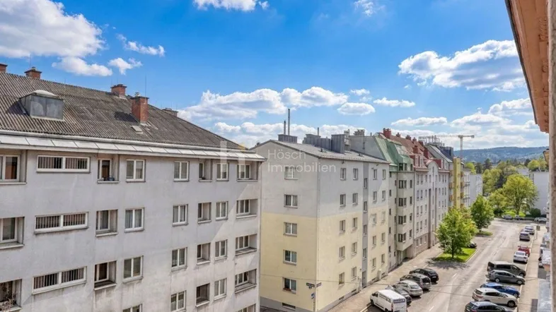 Blick von oben auf eine Wohnstraße mit parkenden Autos und Gebäuden unter blauem Himmel.