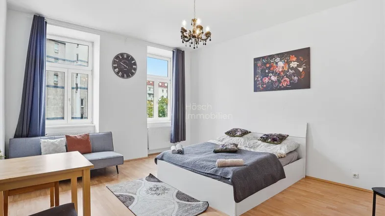 Helles Wohn- und Schlafzimmer mit Holzboden, bequemer Couch und einem Kronleuchter.