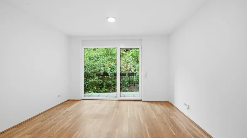 Helles Zimmer mit Holzboden und Zugang zu einem Balkon mit Blick ins Grüne.