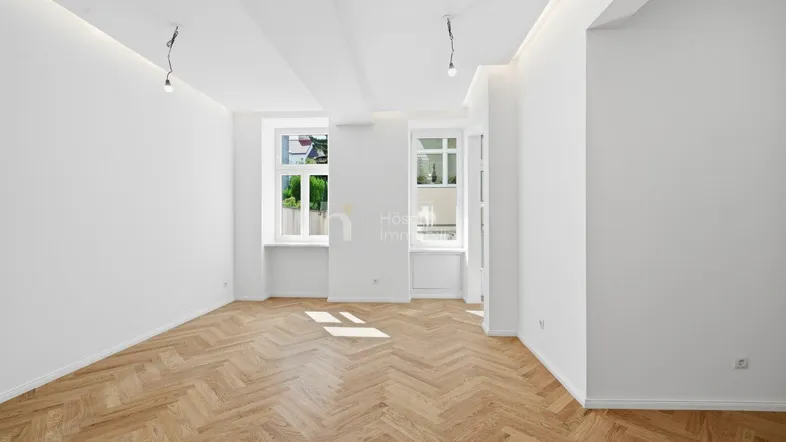 Geräumiges Zimmer mit elegantem Fischgrätparkett und viel Tageslicht durch die Fenster.