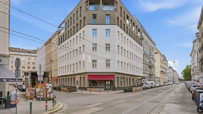 Modernes Eckgebäude in urbaner Umgebung mit Straßenbahnschienen und Geschäften im Erdgeschoss.