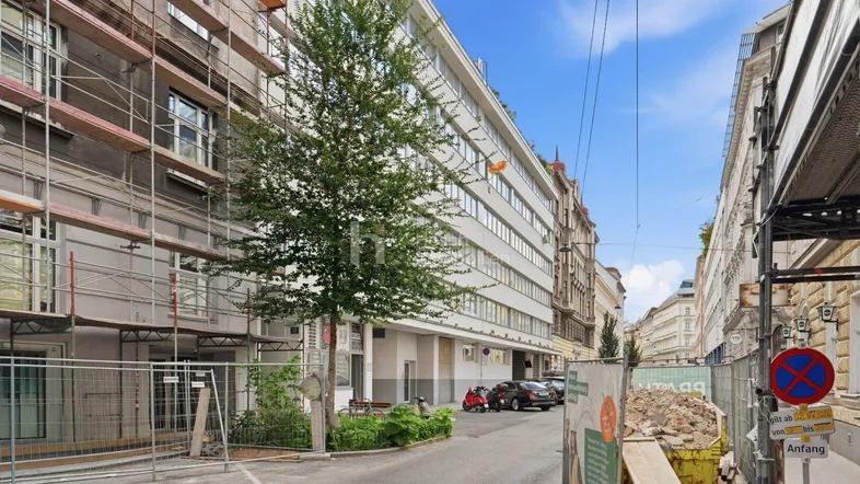 Straßenansicht mit Gebäuden, eines davon im Bau, und parkenden Fahrzeugen in einer städtischen Umgebung.