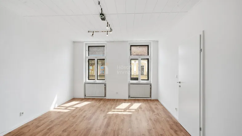 Helles Zimmer mit Laminatboden, weißen Wänden und zwei Fenstern, die viel Tageslicht hereinlassen.