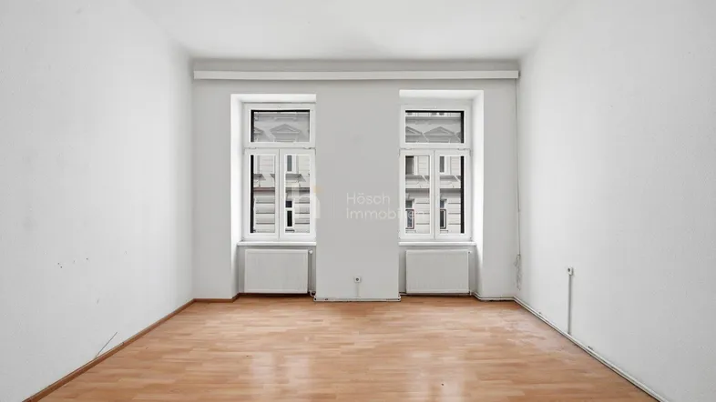 Helles, leeres Zimmer mit zwei Fenstern, Heizkörpern und Laminatboden.