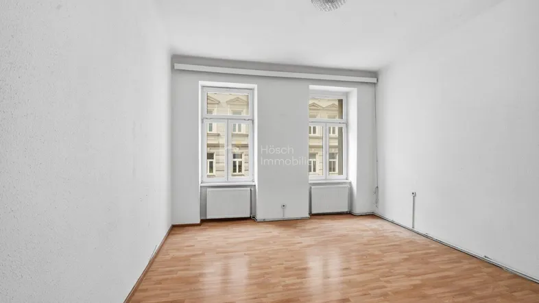 Helles, leeres Zimmer mit Laminatboden und zwei Fenstern, die auf die Straße blicken.