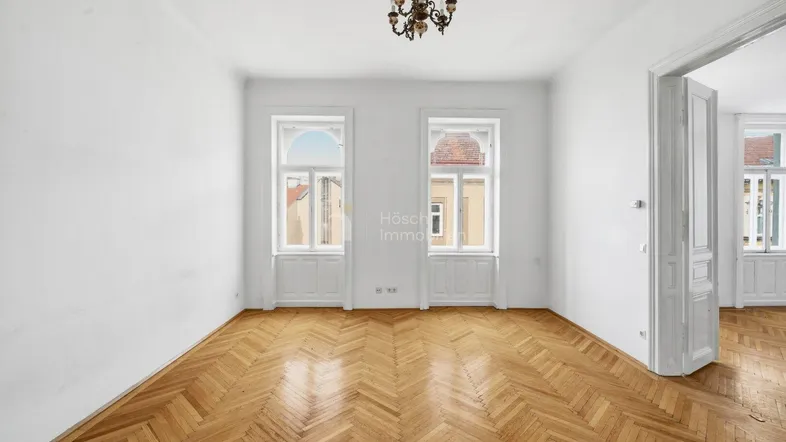 Helles Zimmer mit Fischgrätparkettboden, hohen Fenstern und einem Kronleuchter, ideal als Wohn- oder Schlafzimmer.