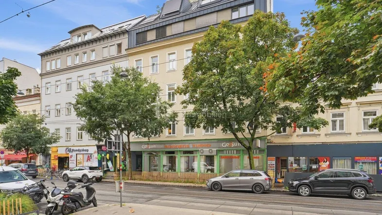 Mehrfamilienhaus mit Geschäften im Erdgeschoss und modernen Dachausbauten an einer belebten Straße.