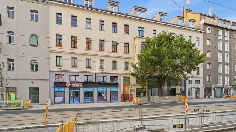 Gebäudefassade mit mehreren Etagen und Geschäften im Erdgeschoss an einer belebten Straße.