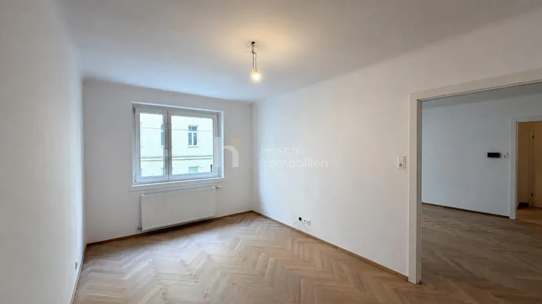 Helles, leeres Zimmer mit Fischgrätparkett und einem Fenster, das viel Tageslicht hereinlässt.
