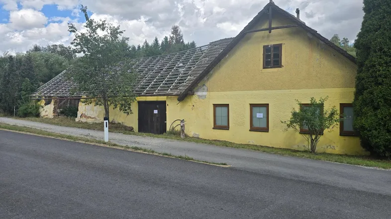 Altes gelbes Bauernhaus mit beschädigtem Dach und grüner Umgebung an einer Straße.