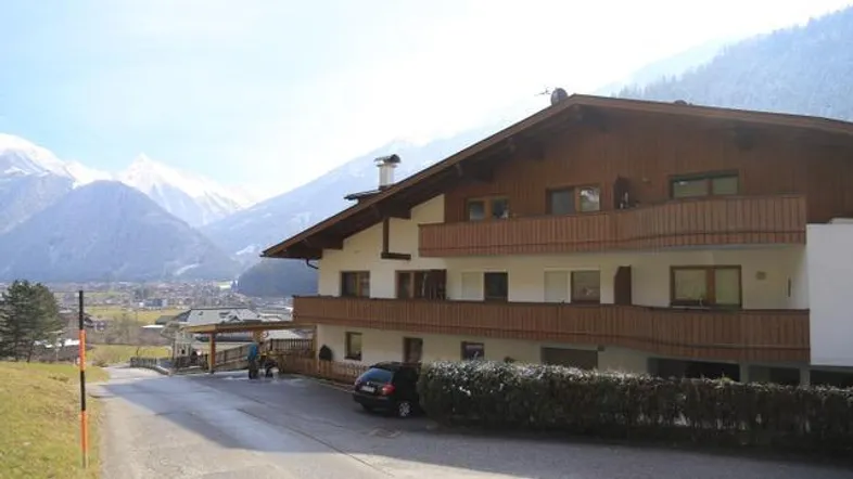 Mehrstöckiges Gebäude mit Holzfassade und Balkonen vor alpiner Bergkulisse und Carport.