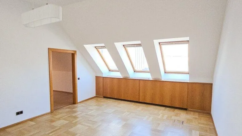 Heller Dachgeschossraum mit schrägem Dach, Parkettboden und drei Dachfenstern, die viel Tageslicht hereinlassen.