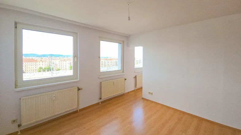 Helles, leeres Wohnzimmer mit großen Fenstern, Parkettboden und Heizkörpern, bietet Weitblick.