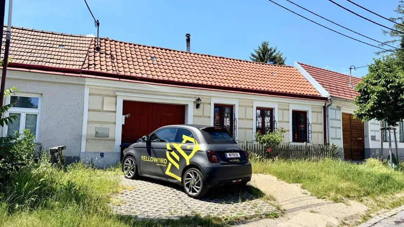 Hausfassade mit rotem Ziegeldach und einer Einfahrt, umgeben von Grünflächen.