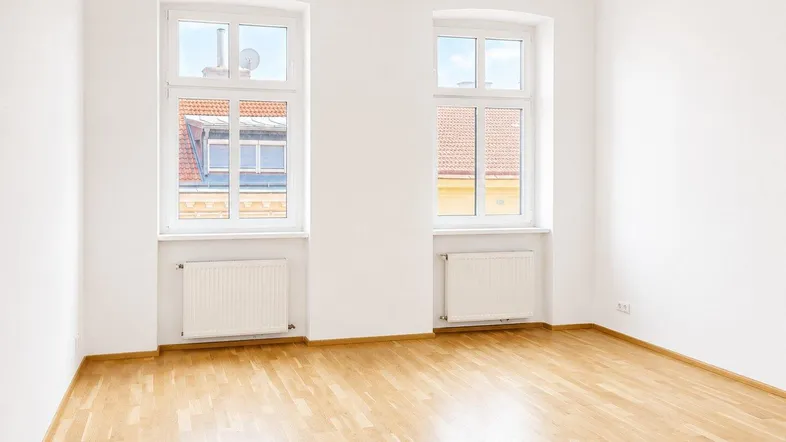 Helles Zimmer mit zwei Fenstern, Holzboden und weißen Wänden, ideal als Wohn- oder Schlafzimmer.