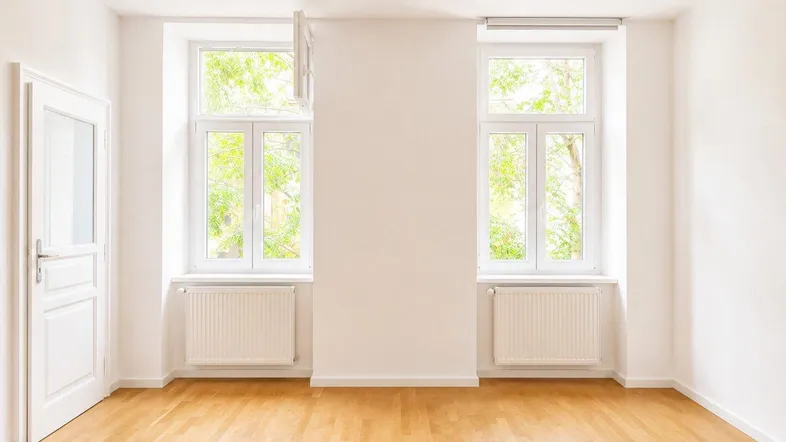 Helles, leeres Zimmer mit Parkettboden und zwei großen Fenstern, die viel Tageslicht hereinlassen.