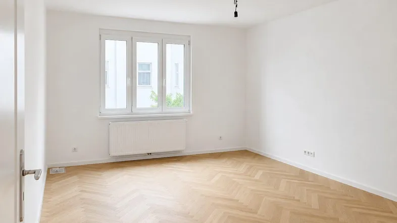 Geräumiges, unmöbliertes Zimmer mit hellem Fischgrätparkett und Fenster, ideal als Schlafzimmer.