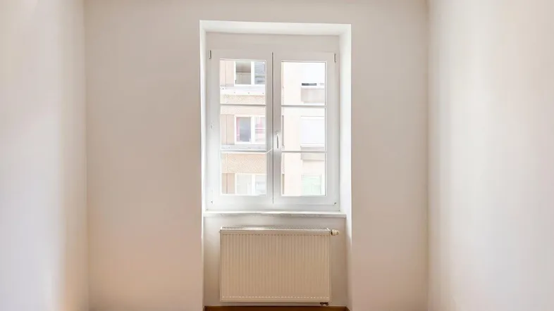 Freundliches Zimmer mit großem Fenster, Holzboden und weiß gestrichenen Wänden.