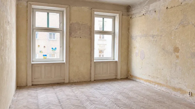 Unrenoviertes Zimmer mit Fischgrätparkett und zwei hohen Fenstern, bereit zur Sanierung.