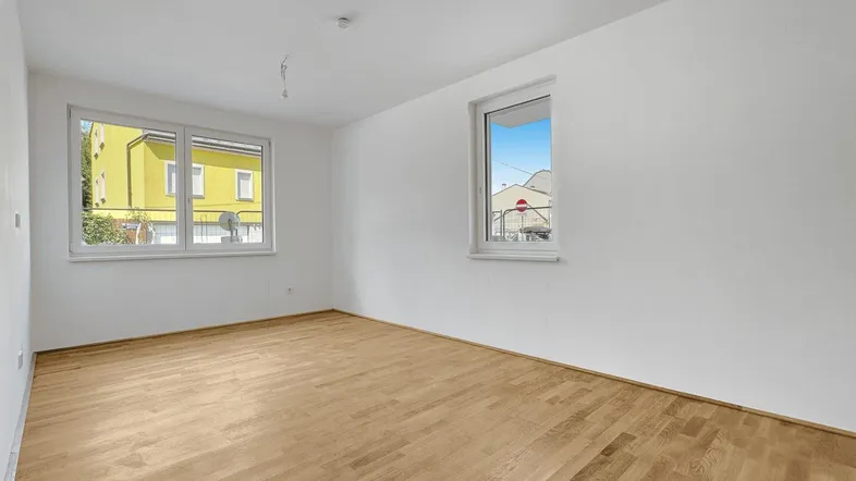 Helles Zimmer mit weiß gestrichenen Wänden und hellem Parkettboden, mit Blick auf ein Nachbargebäude.