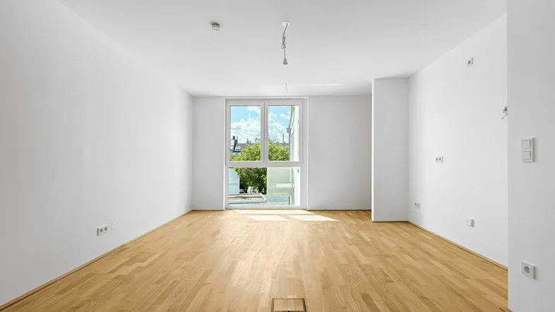 Heller, leerer Wohnraum mit neuem Parkettboden und großen Fenstern, die viel Tageslicht hereinlassen.