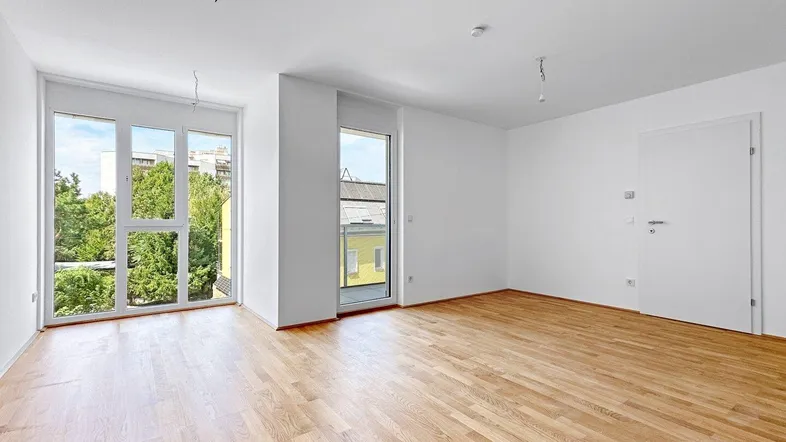 Helles, leeres Wohnzimmer mit großen Fenstern, Holzboden und Blick ins Grüne.